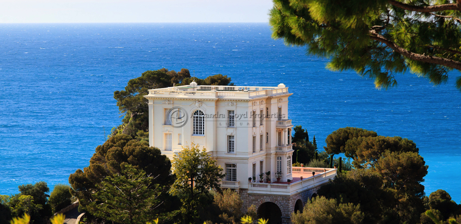 A large white building sits on a hill, overlooking a serene ocean