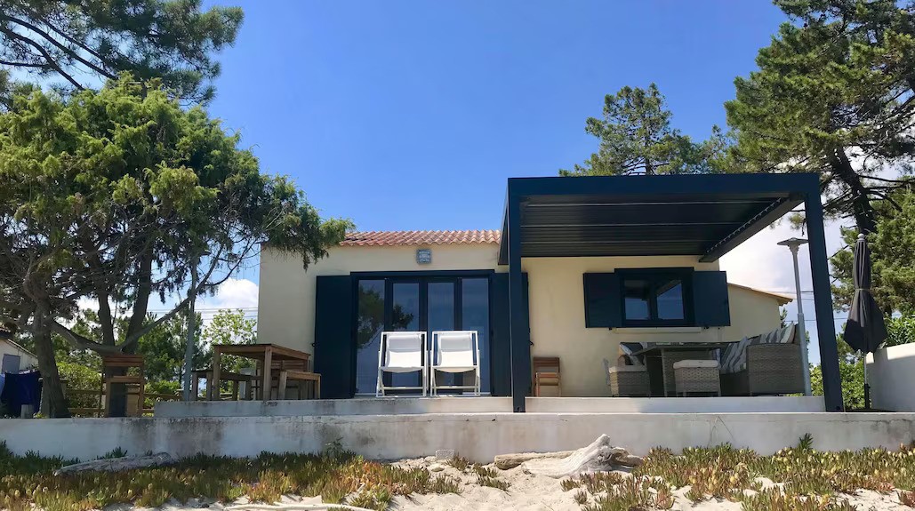 A small beach house with a patio, surrounded by sand and ocean waves